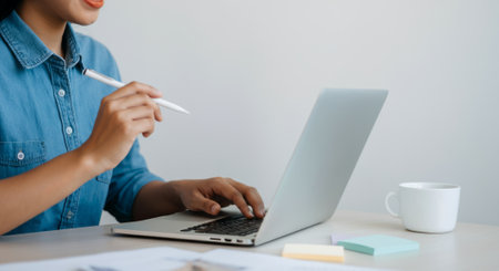 Woman is typing on a laptop computer. She is holding a pen in her hand. There is a cup on the table next to herの素材