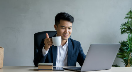 Man is sitting at a desk with a laptop and a cup of coffee. He is smiling and holding the cupの素材