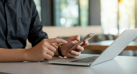 Man is using a cell phone and laptop at a table. He is pointing at the cell phoneの素材