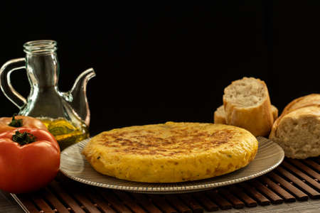 Spanish tortilla on a rustic plate and a wooden tablecloth with a glass jar of olive oil, some...