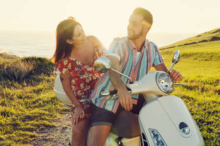 Couple of young friends with fresh clothes, having fun and smiling on a motorcycle in the coast during the sunset lightの写真素材