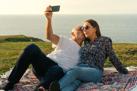 Aged Couple laying on the meadow, taking some selfies and enjoying the sunset on the coastの写真素材
