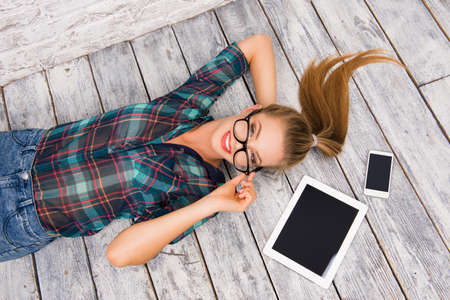 Top view of cute smart young woman lying on wood floor with tablet, glasses and phoneの写真素材