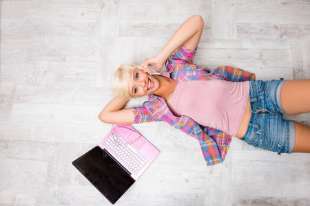 Top view of young woman lying on floor with laptop and talking on phoneの写真素材