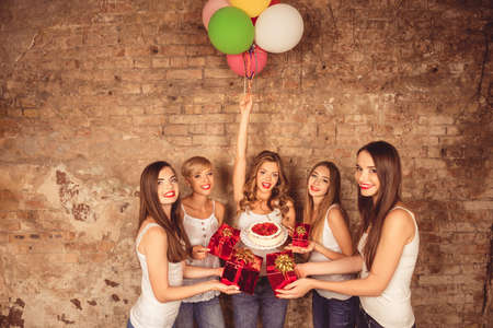 Cheerful pretty girls holding cake and balloons giving presents to their friendの写真素材