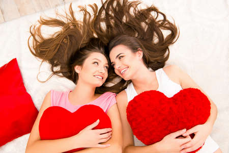 Cheerful beautiful girls in pajamas lying on the bed holding pillowsの写真素材