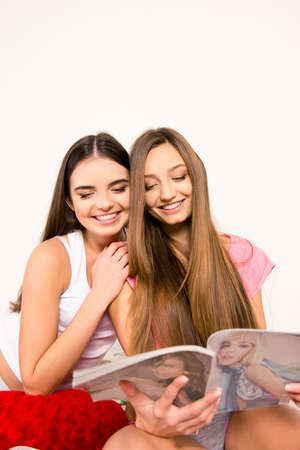 Cute cheerful girls in pajamas sitting on the bed reading magazinesの写真素材