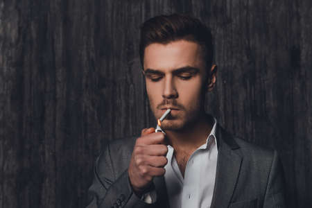 Handome brutal man in suit on the grey background lighting a cigaretteの写真素材