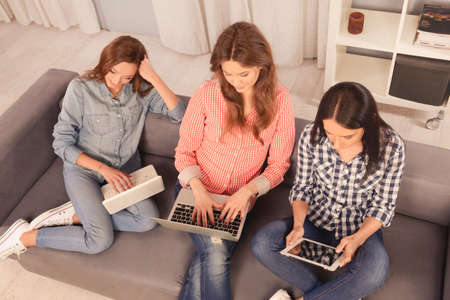 Three cute  girls sitting on the couch and working on laptopの写真素材