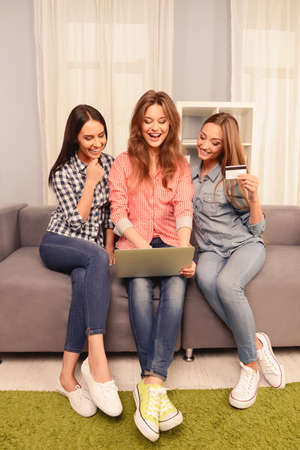 Three laughing women sitting on sofa with laptop and making purchacesの写真素材