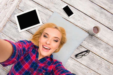 Cheerful blonde with tablet, phone, cup and glasses making selfie