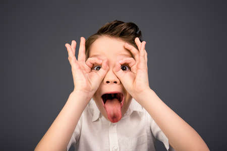 Comic little boy holding hands like glasses and showing tongue