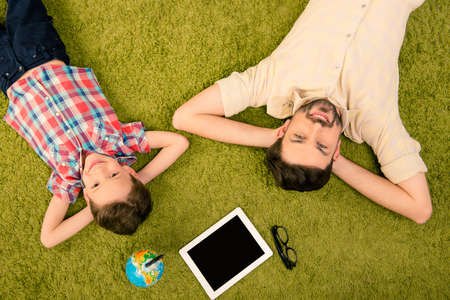 Top view of father and son lying on green carpet with globe, tablet and glassesの写真素材