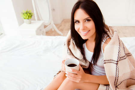 Smiling happy woman basking under plaid with cup of hot teaの写真素材