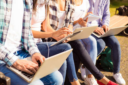 close-up photo of happy diverse classmates sitting on bench and study up with deviceの写真素材