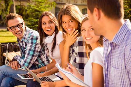 five smiling happy beautiful classmates talking and sitting on bench with device