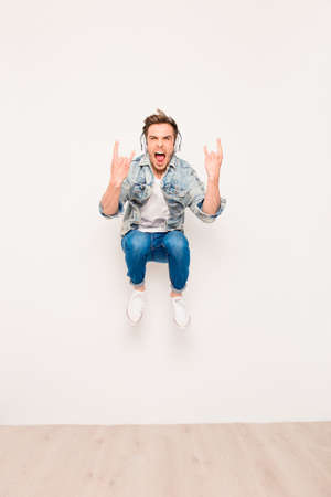 Crazy young man listening music in headphones, jumping and showing rock gesture