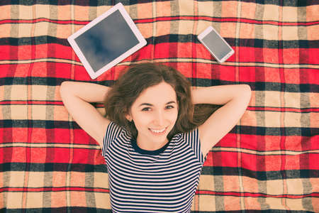 Pretty happy girl lying on plaid with digital tablet and smartphoneの写真素材