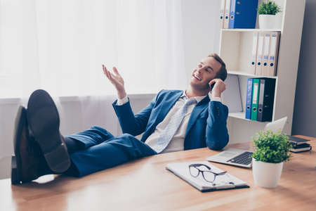 Happy businessman talking on phone and holding legs on tableの写真素材