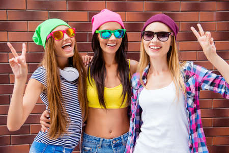 Cheerful girlfriends in hats and glasses gesturing with two fingersの写真素材