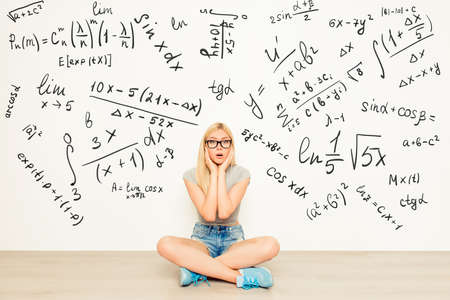 attractive shocked girl in spectacles  sitting on floor near wall with math formulas, crossed legs and touching faceの写真素材