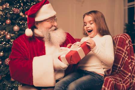 wow dream is real in christmastime! happy little cute girl sitting on the lap of aged Santa Claus. He giving red  box with bow of ribbon for she at home near christmas treeの写真素材