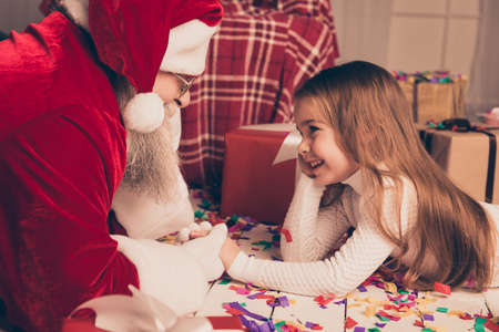 it's christmastime emotion!  Happy Santa Claus wearing red costume lying on floor with cute little girl, holding hands and having funの写真素材