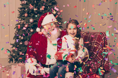 it's christmastime! At party with confetti Santa Claus wearing red costume sitting on armchair in living decorated room with cute little girl holding gift with bow and having funの写真素材