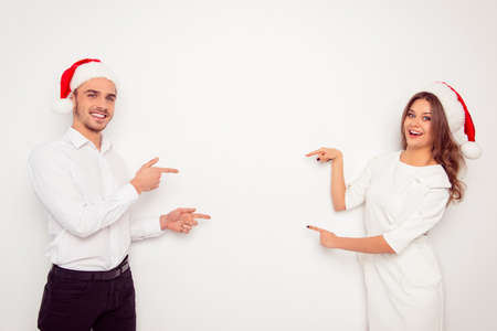 Happy cheerful man and woman in red santa hats pointing on each otherの写真素材