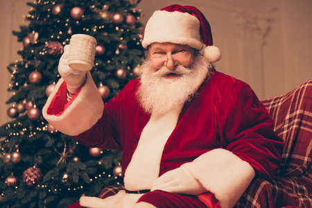 Cheerful santa claus in red costume sitting near pine and holding cup of teaの写真素材