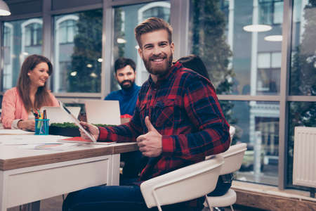 Young handsome man showing thumb up while his collegues working on project, sitting on modern workplaceの写真素材