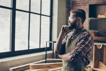successful handsome cabinetmaker look to the side at window. Stylish young entrepreneur with brutal hairstyle and glasses holding chin and dream at his workstationの写真素材
