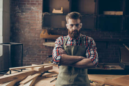 serious successful handsome joiner look at camera near tabletop with tools.  Stylish young entrepreneur with brutal hairstyle and saved glasses crossed hands at his workstationの写真素材