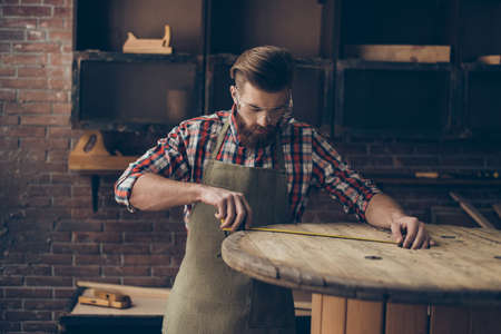 serious handsome  craftsman work with ruler. Stylish young cabinet-maker with brutal hairstyle and saved glasses work at his workplaceの写真素材