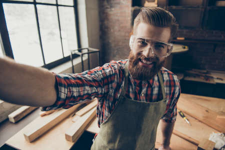 Happy successful handsome cabinetmaker make selfie photo. Stylish young entrepreneur with brutal hairstyle and glasses smiling and photographing at his workstation. Love jobの写真素材