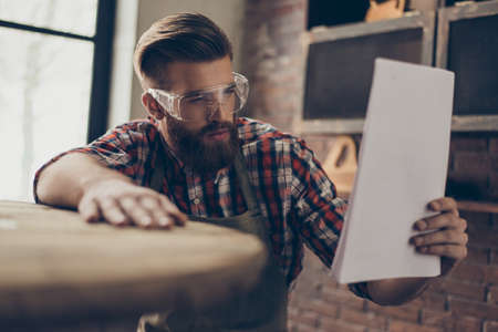 serious handsome  craftsman chek wood and document. Stylish young cabinet-maker with brutal hairstyle and saved glasses work at his workplace. He love his jobの写真素材