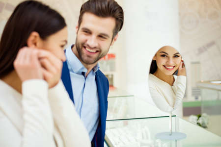 Happy beautiful young woman with her success boyfriend trying earring near mirror in jewelry storeの写真素材