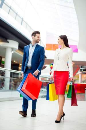 Portrait of happy lovely cute young woman with her handsome boyfriend in suit  holding colored shopping packages and together walk  in modern mall and having fun.の写真素材