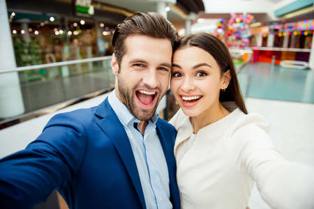 Happy lovely young woman with her handsome boyfriend in suit together make selfie photo, walk in modern mall and having fun.の写真素材