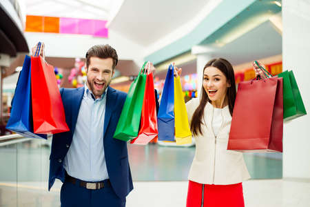 Photo of cheerful stylish successful happy young lovely couple raised  colored shopping bags and laughing in mall. Concept of consumerism, sale, rich life and people relationship.の写真素材