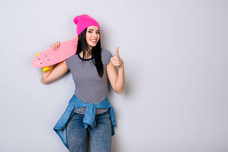 Young girl in pink hat and skateboard on her shoulder smiling and showing thumb upの写真素材