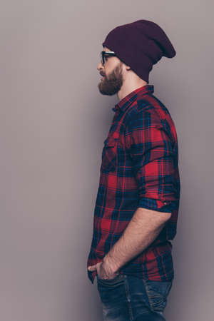 Young stylish guy in jeans, checkered shirt and glasses with rolled up sleeves standing in profile and holding his hands in pocketsの写真素材