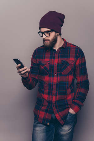 A vertical portrait of young bearded guy in checkered shirt and hat tyling sms and holding his hand in  pocket against gray backgroundの写真素材