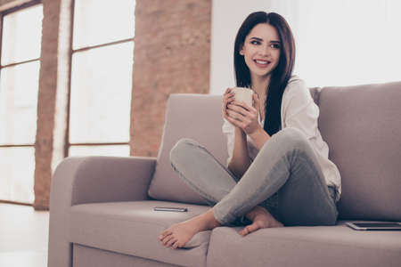 Portrait of beautiful young woman sitting on sofa at home with cup gf tea near big window, crossing...