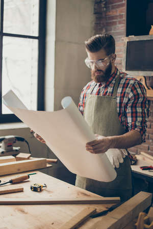 Stylish young woodwoker with glasses in checkered shirt reading blueprint at his workshop.の写真素材