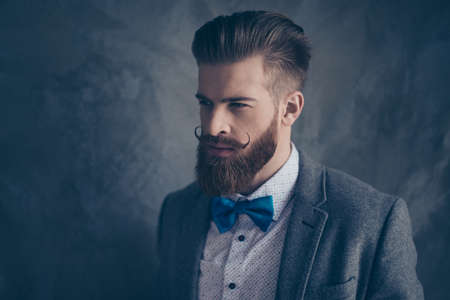 Portrait of stylish hipster brutal young man with red mustache, beard and beautiful hairstyle ready for event party while standing on gray backgroundの写真素材