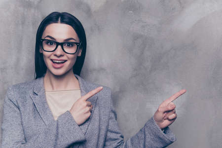 Portrait of excited cheerful young successful businesswoman in glasses and suit pointing to the side  while standing near gray wallの写真素材