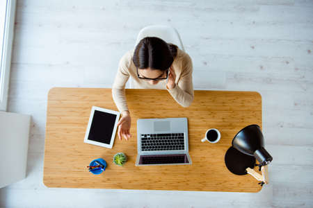 Top-view of young successful woman in glasses sitting at table with laptop, cup of coffee and tablet...