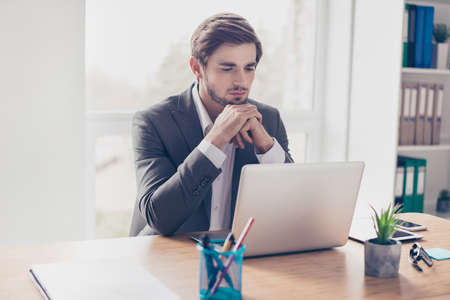 young handsome serious man is sitting at  office desk in workstation and looking thoughtfully at laptop screenの写真素材
