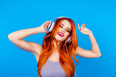 Carefree happy joyful girl with beautiful red curly long hair listen to music with white headphones  while standing on blue backgroundの写真素材